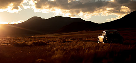 A Porsche sports car is parked in a field with mountains in the background at sunset.