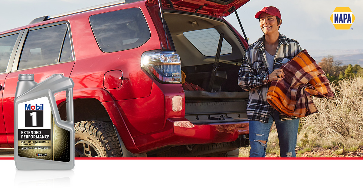 A woman is outside at the trunk of her car holding a premium travel blanket, a Mobil 1 motor oil bottle is in the foreground.